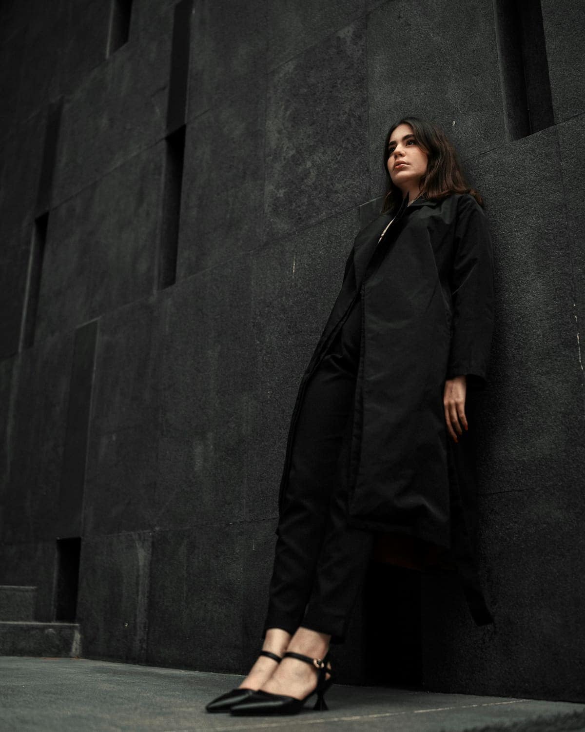 Model in a long black coat against a dark stone wall